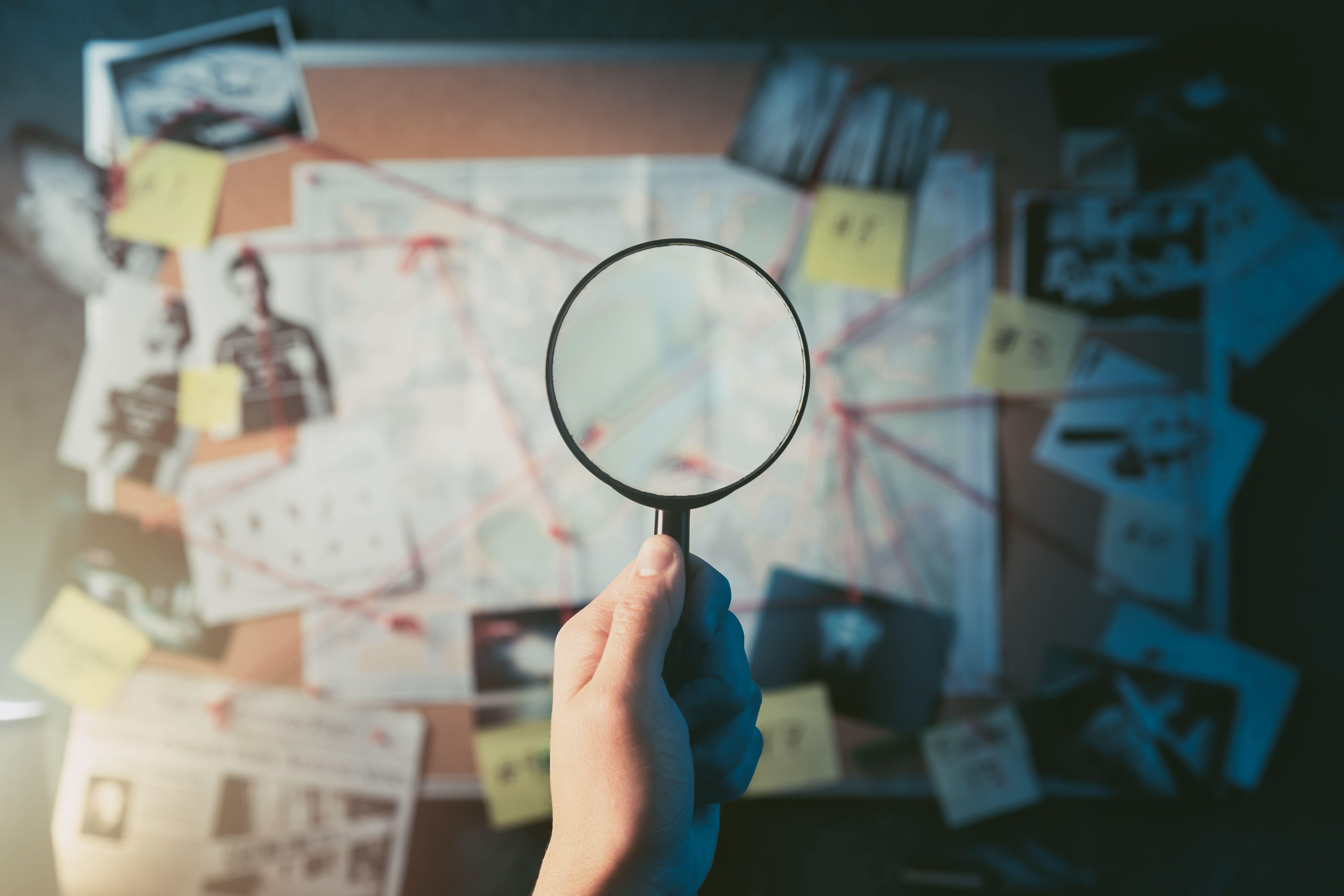 Detective Hand Holding a Magnifying Glass in Front of a Board With Evidence